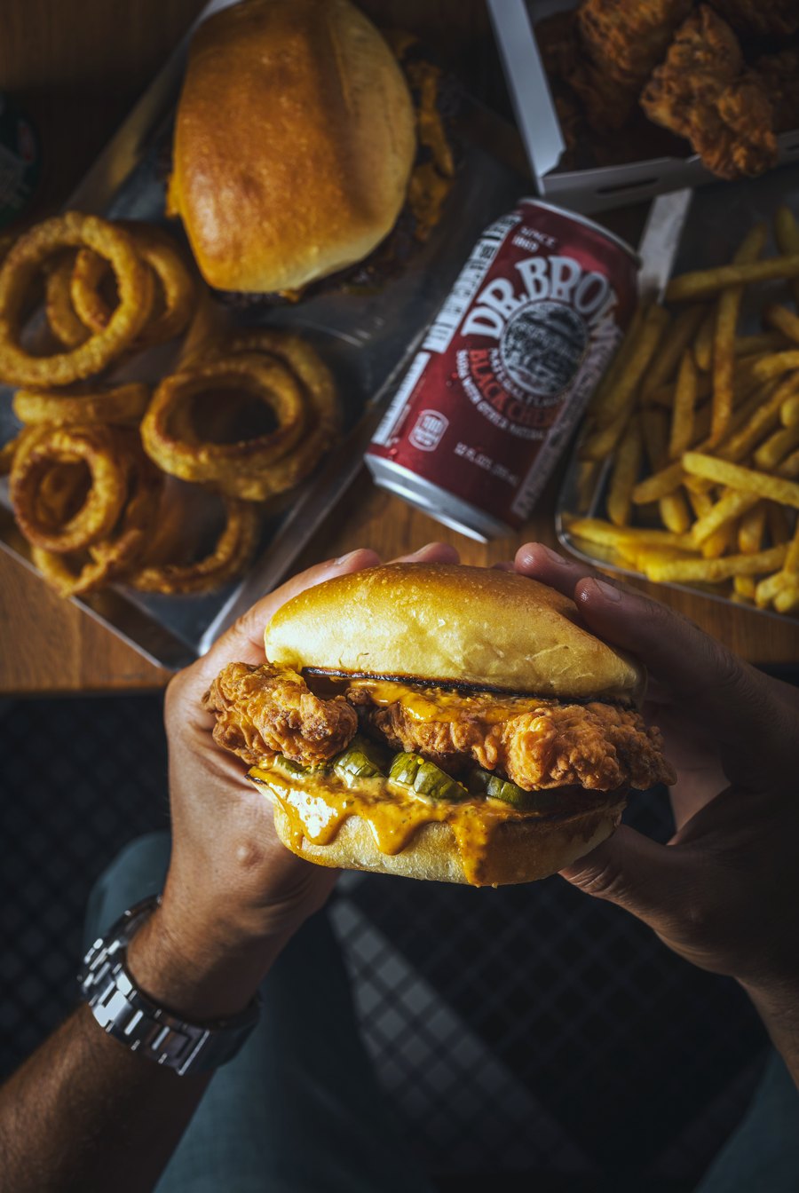 Crispy chicken sandwich in hand, with onion rings, fries and Dr. Brown's